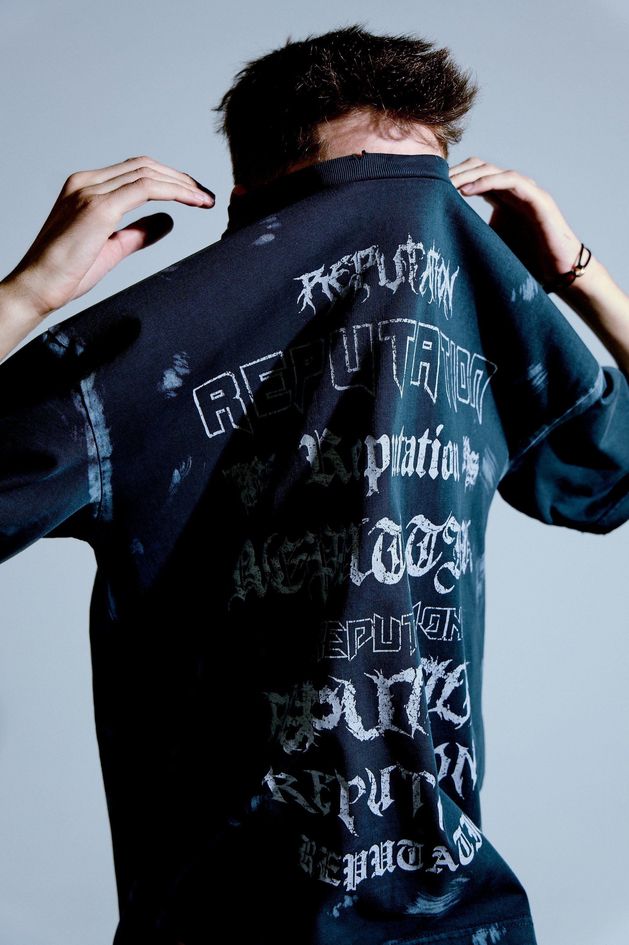 Asit yıkamalı koyu tee’nin arka yüzünde çoklu “Reputation” tipografi baskısı; yarım portre. / Acid-washed dark tee with multi “Reputation” typography back print; half-portrait.