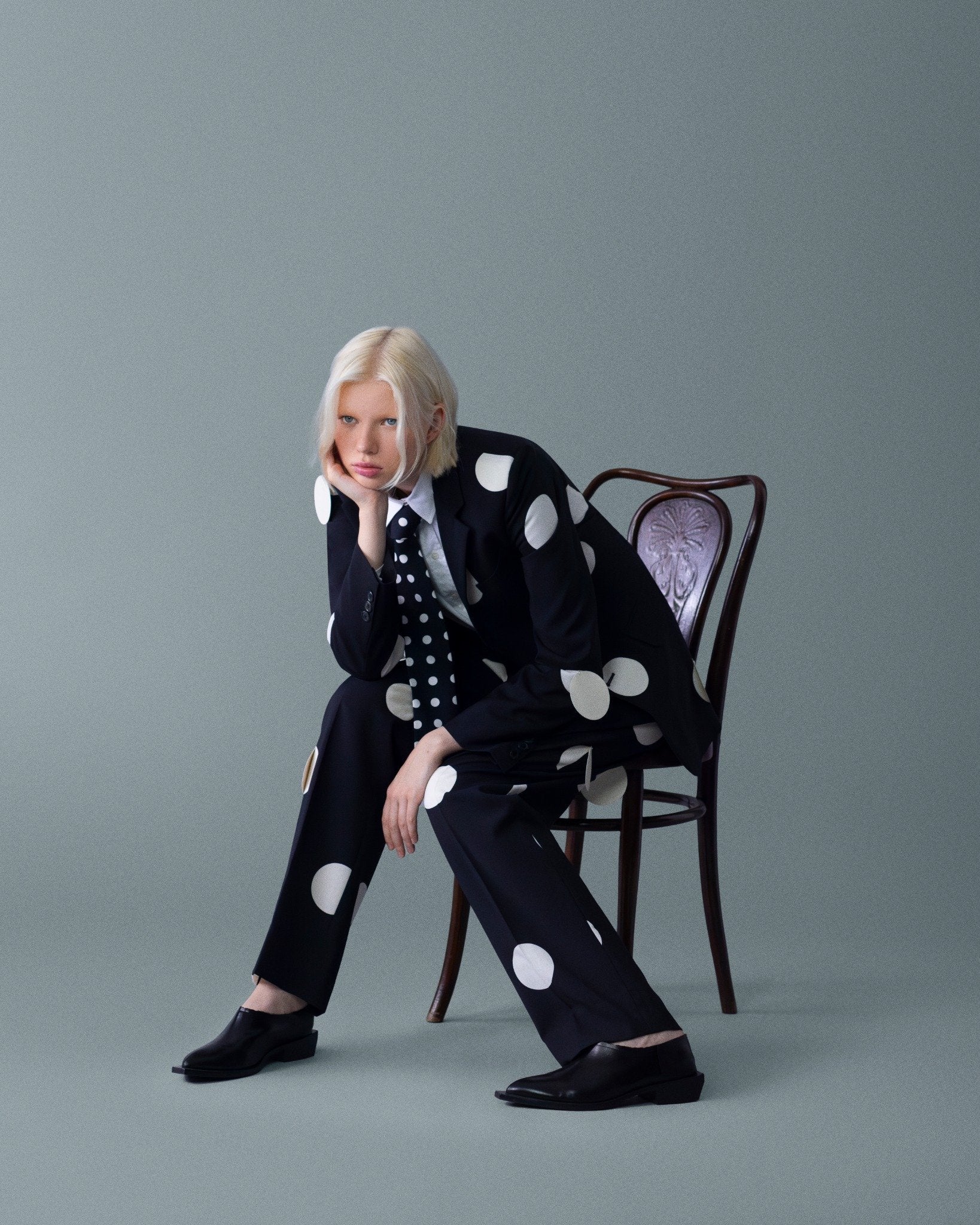 Büyük puantiyeli siyah takım ve noktalı kravat; sandalyede cool, androjen bir tavır. \ Black suit with oversized polka dots and a dotted tie; a cool, androgynous attitude on a chair.
