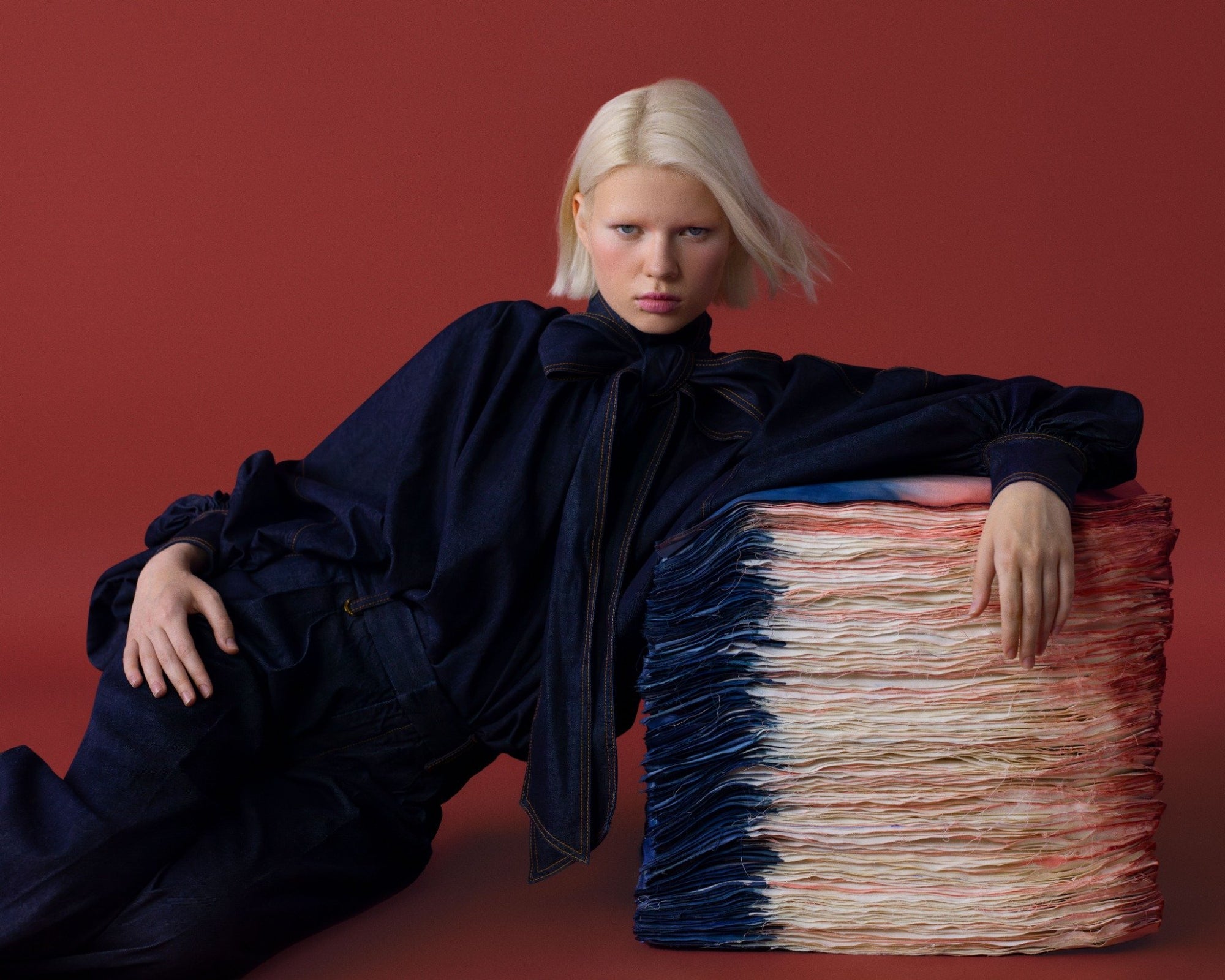 Koyu denim fiyonk yaka bluz ve indigo takımla sanat nesnesine yaslanan güçlü kadraj; tuğla kırmızısı fon ile yoğun kontrast. \ Dark-denim bow-neck blouse and indigo set leaning against an art object; bold contrast with a brick-red backdrop.