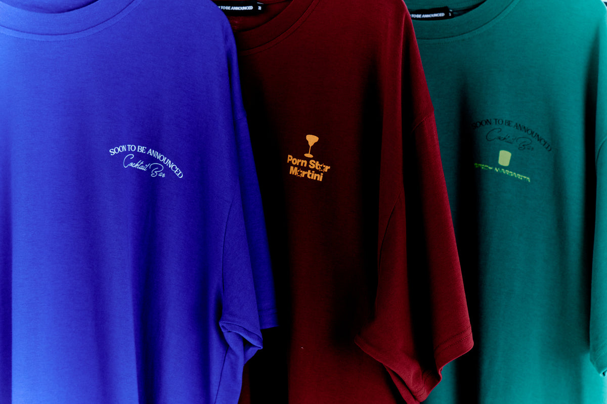 Soontobeannounced “Cocktail Bar” serisi tişörtler; mor, bordo ve teal renklerde, göğüste kokteyl temalı nakış/print logolarla, askıda stüdyo çekimi.

Soontobeannounced “Cocktail Bar” T-shirts in purple, burgundy and teal with cocktail-themed chest embroidery/prints; studio shot on hangers.