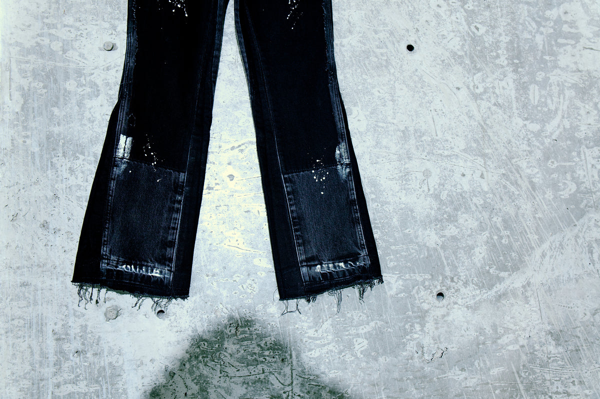 Soontobeannounced siyah denim pantolon paça detayı; paint-splatter efektli, püsküllü paça, dokulu beton zemin üzerinde stüdyo çekimi.

Soontobeannounced black denim trousers hem detail with paint-splatter and raw hems; studio shot on a textured concrete floor.