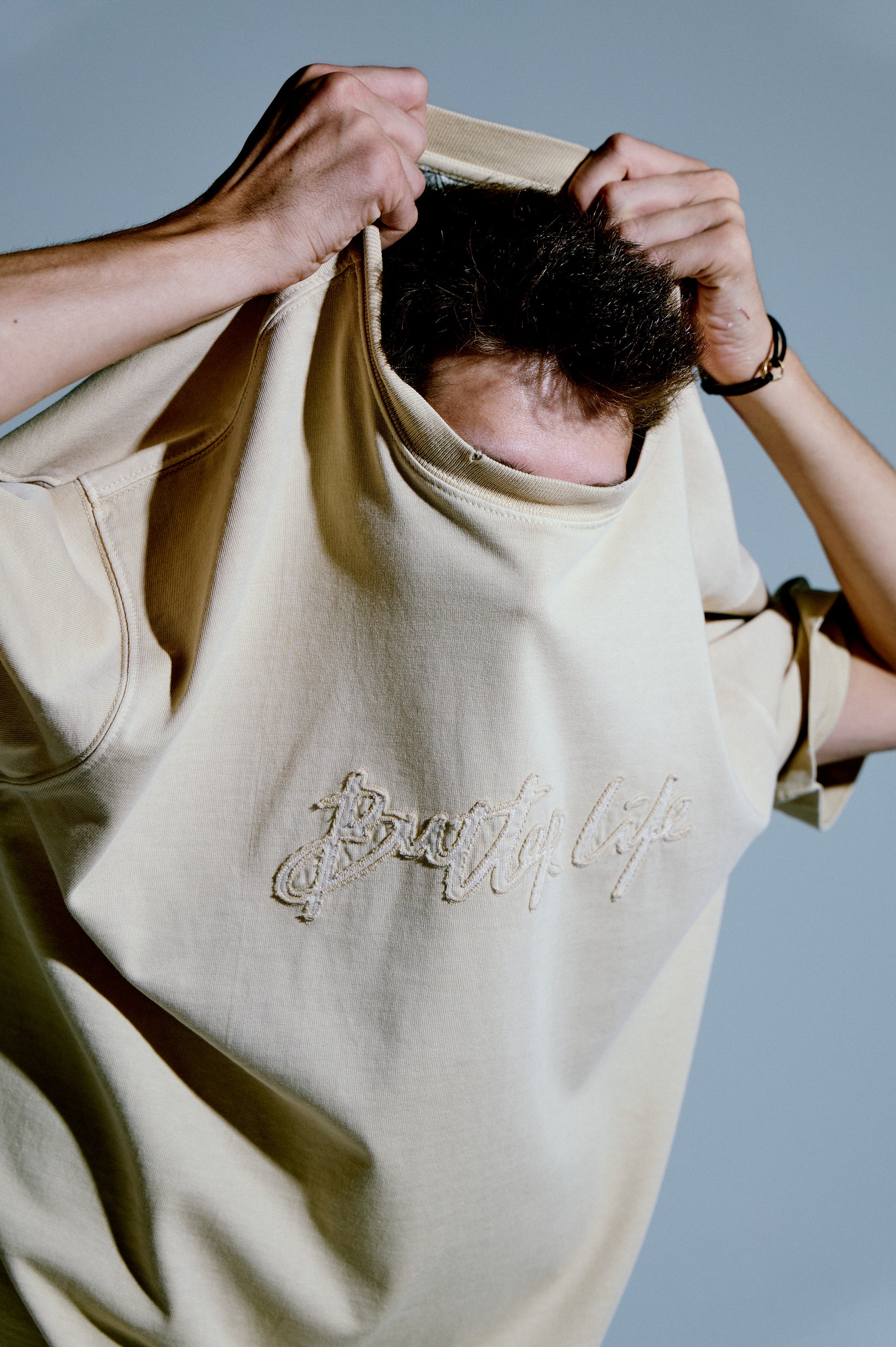 Soontobeannounced bej tişört; göğüste işleme yazı detaylı, model tişörtü başından geçirirken stüdyo ışığında yakın plan.

Soontobeannounced beige T-shirt with embroidered script logo; close-up of a model pulling the shirt over the head under studio light.