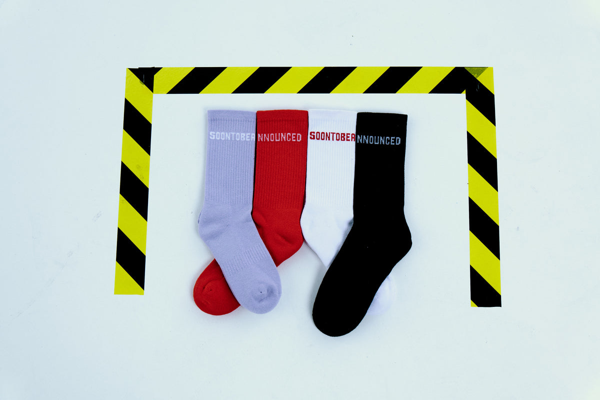 Soontobeannounced logolu kırmızı, lila, beyaz ve siyah çoraplar; stüdyo çekimi, sarı-siyah şerit çerçeve üzerinde.

Soontobeannounced logo socks in red, lilac, white and black; studio shot on a yellow-black striped frame.
