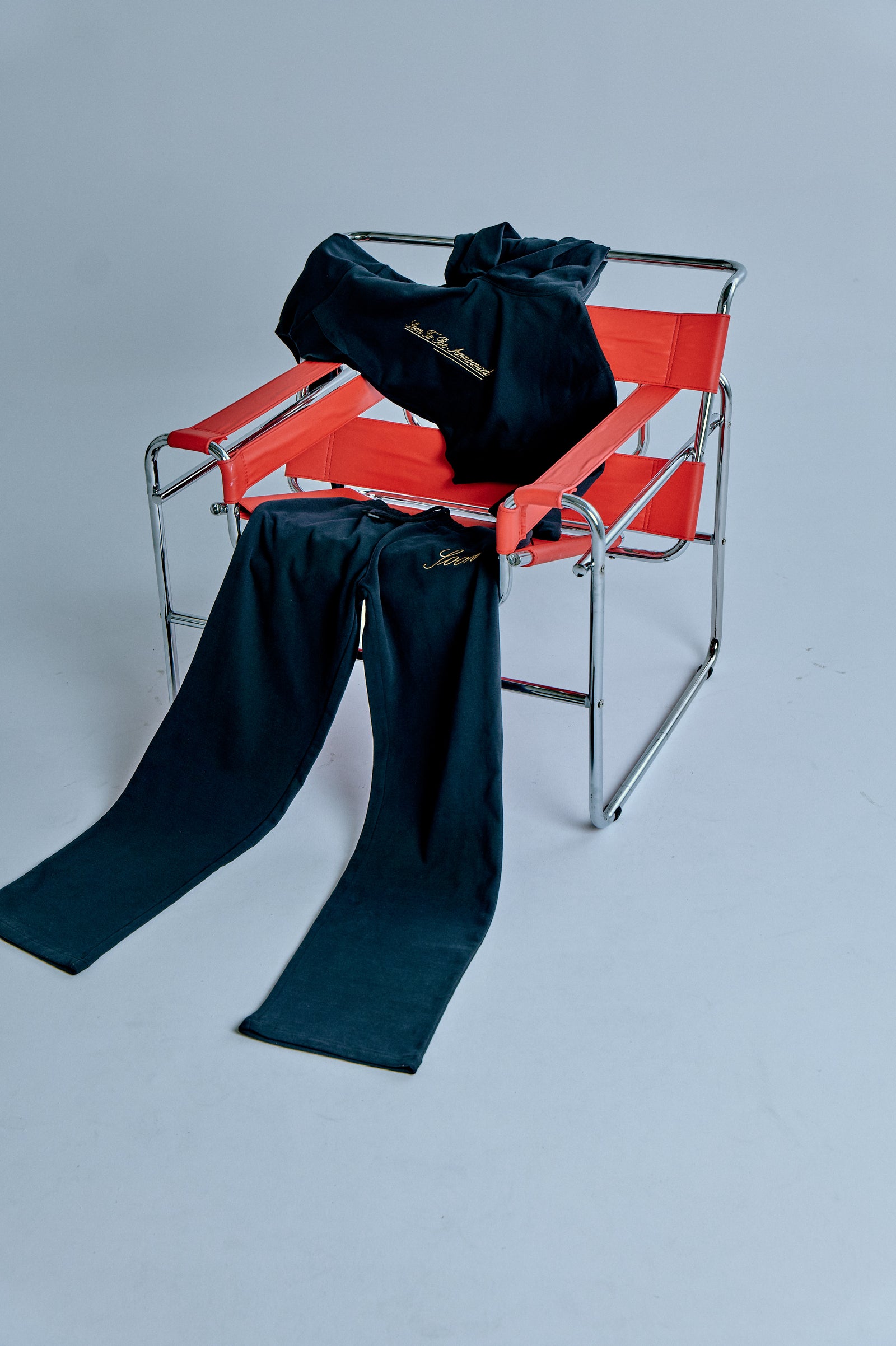 Kırmızı sandalyeye serilmiş eşofman takım; minimal kompozisyonda üst ve paçalar vurgulu stüdyo planı. / Tracksuit draped over a red chair; minimal composition highlighting hoodie and legs — studio shot.