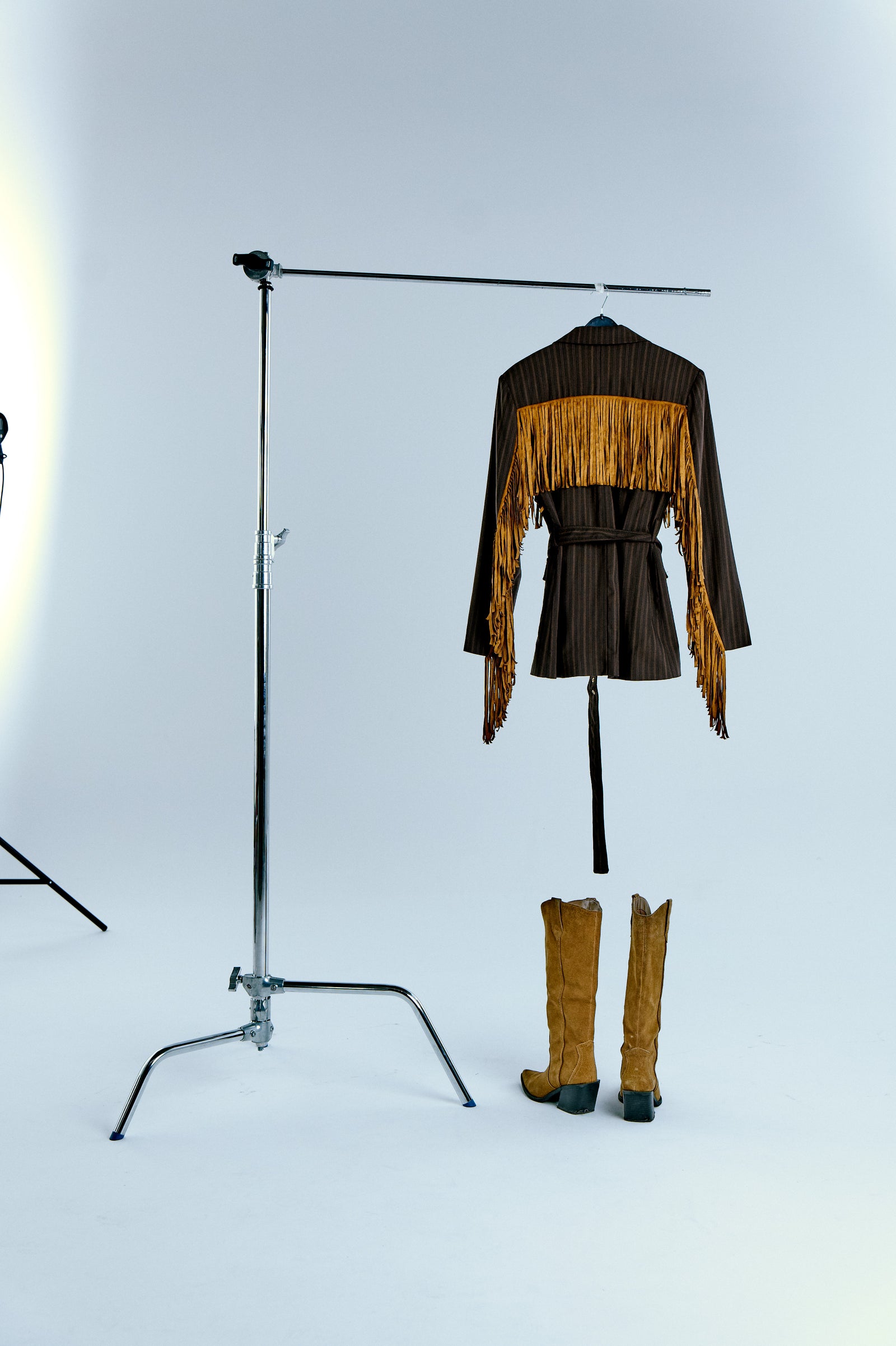 Çizgili blazerin arka görünümü; omuzdan bele uzanan süet püskül detayları ve kovboy çizmeleriyle tam kadraj stüdyo çekimi. / Back view of pinstripe blazer showing long suede fringes; full-frame studio shot with cowboy boots.