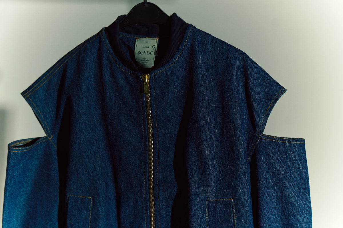 Sorbe denim ceket; kol oyuklu (cut-out) tasarım, kontrast sarı dikişler ve metal fermuar, stüdyo ışığında askıda yakın plan.

Sorbe denim jacket with shoulder cut-out design, contrast yellow stitching and metal zipper; close-up on hanger under studio light.