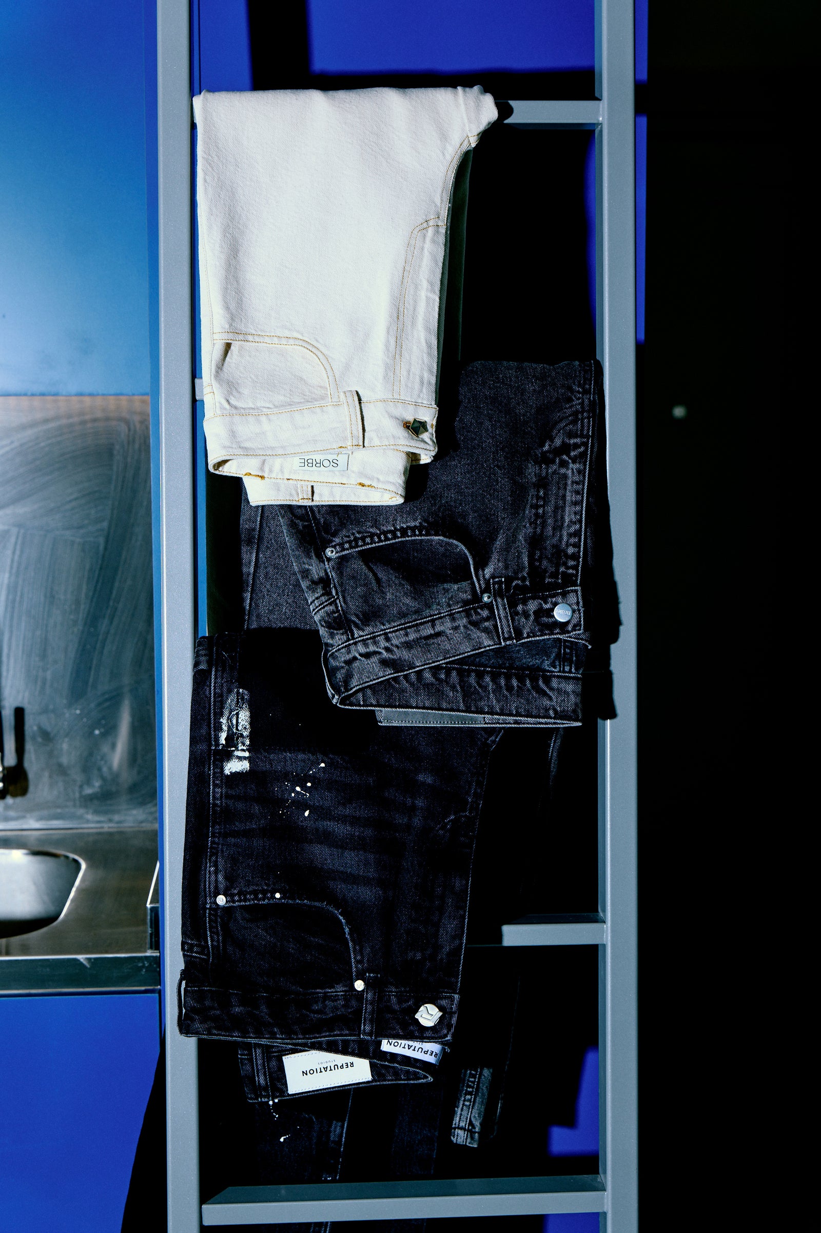 Krem Sorbe jean üstte, altında koyu tonlar; alt parçada boyalı efektli siyah denim vurgusu. / Cream Sorbe jeans on top with darker pairs beneath; lower piece highlighted with paint-splatter effect.