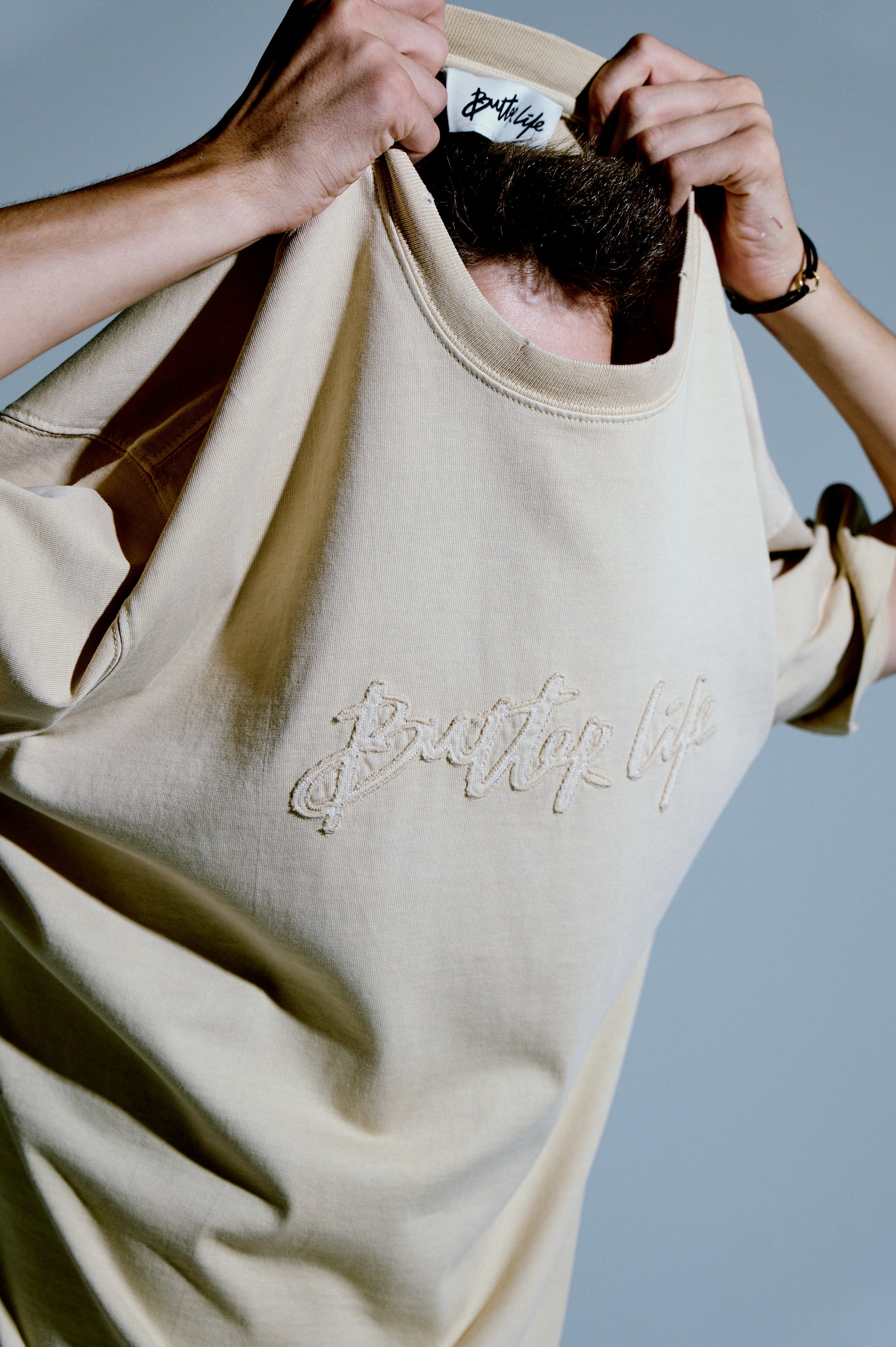 Bej tee; iç yaka etiketi ve “Better Life” nakışı görünür şekilde hareketli kadraj. / Beige tee showing inner neck label and “Better Life” embroidery in a dynamic frame.