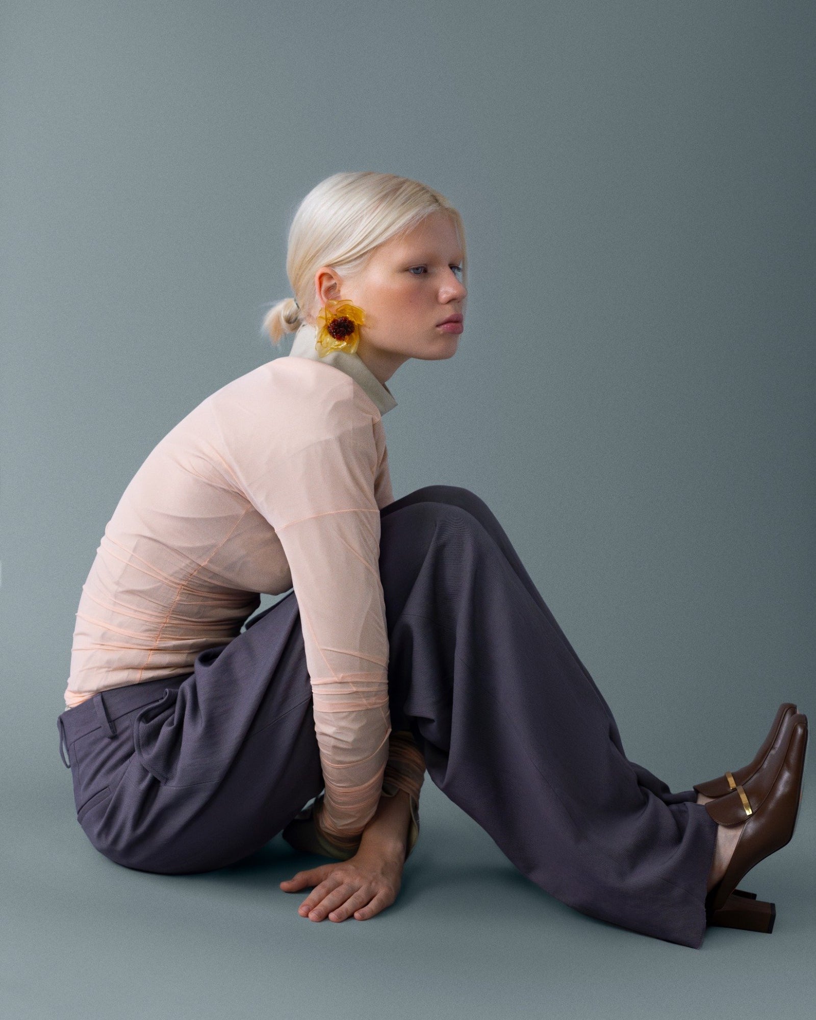 Pastel şeftali üst ve kömür gri paçalarla yere yakın oturuş; kulakta tek güneşçiçek aksesuarı ile şiirsel bir duruş. \ Soft peach top and charcoal wide-legs in a grounded pose; a single sunflower earring adds a poetic note.