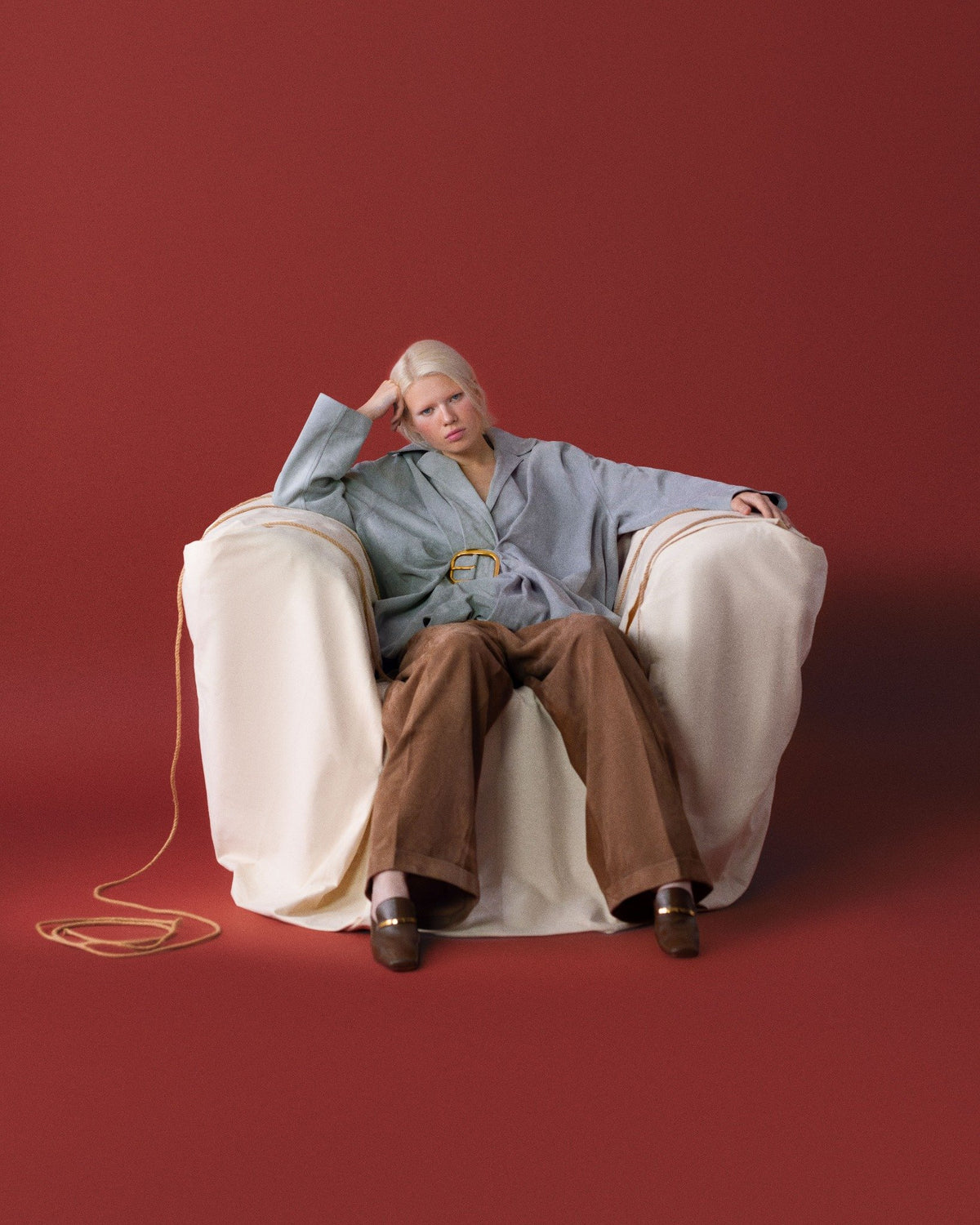 Oversize gri ceket ve kahverengi pantolonla sarılı koltukta rahat bir moda anı; altın tokayla sofistike vurgusu. \ A relaxed fashion moment in an over-sized gray jacket and brown trousers on a wrapped armchair; a gold buckle adds sophistication.