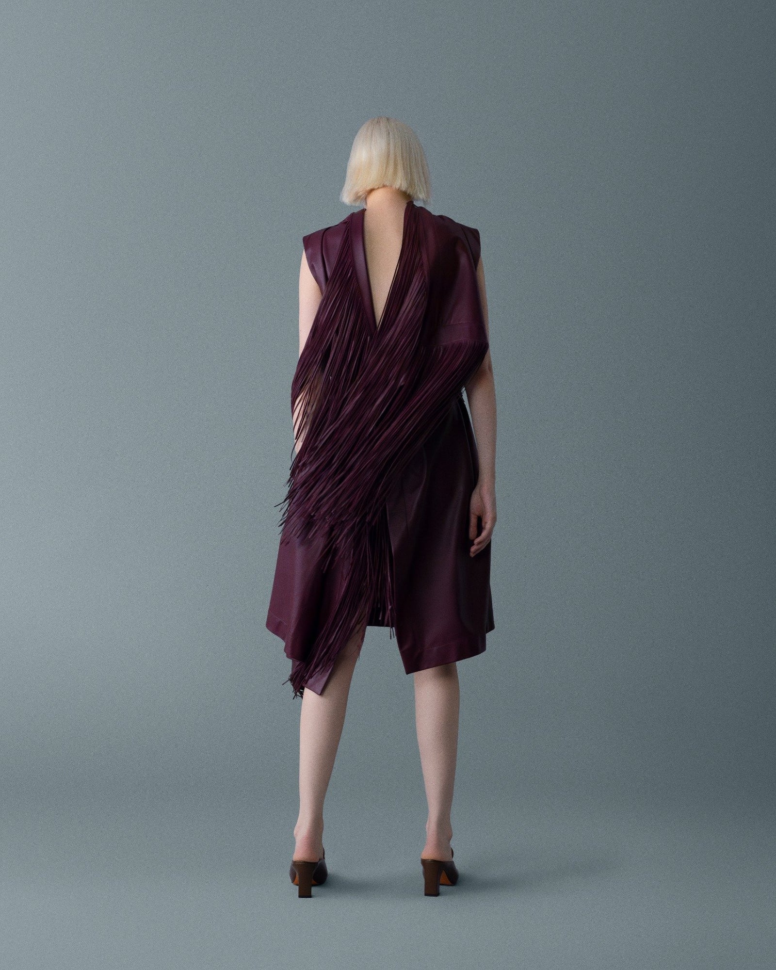 Sırtı derin oyuk, şarap tonlarında, hareketli püskül paneliyle heykelsi bir elbise. Sessiz gri arka planda akış ve doku vurgulanıyor. \ A sculptural wine-toned dress with a deep back and a cascading fringe panel. Flow and texture take center stage against a quiet gray backdrop.